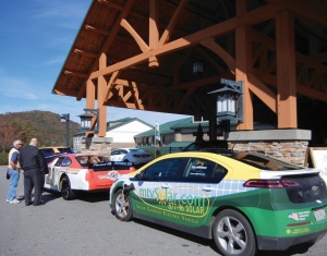 Two cars with marketing decals on display at an event while two men speak in the background.